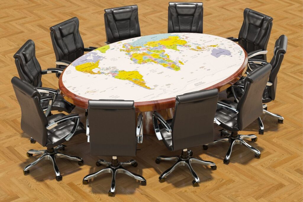 Uma mesa de reunião oval com um mapa mundi como tampo, cercada por cadeiras de escritório pretas, simbolizando diplomacia e governança global.