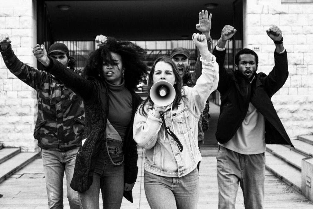 Grupo de jovens adultos de diversas etnias em preto e branco, com punhos erguidos e uma mulher no centro segurando um megafone, expressando protesto e engajamento político.