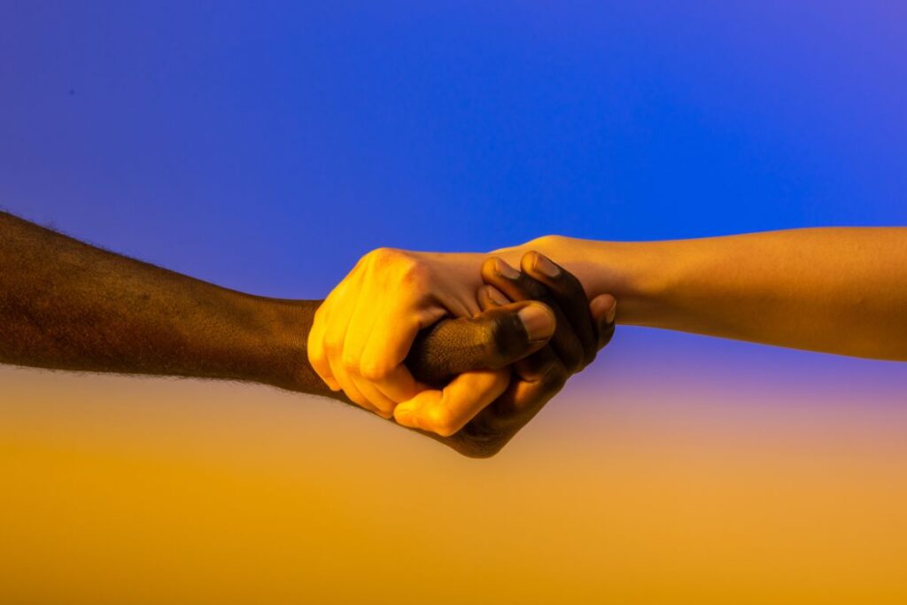 Mãos de pele clara e escura se apertando contra um céu gradiente de azul para amarelo, simbolizando união, diversidade e cooperação na cultura política.
