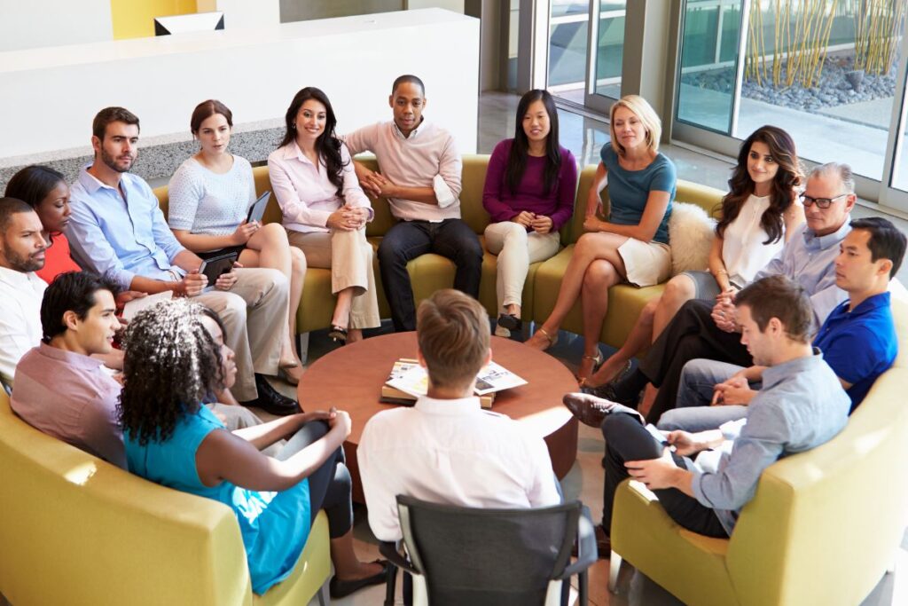 Grupo diversificado de pessoas sentadas em um círculo em poltronas verdes, com uma mesa redonda no centro, simbolizando diálogo e participação em decisões.